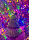 Decorative glass Christmas tree held in front of a lit Christmas tree with colorful lights.